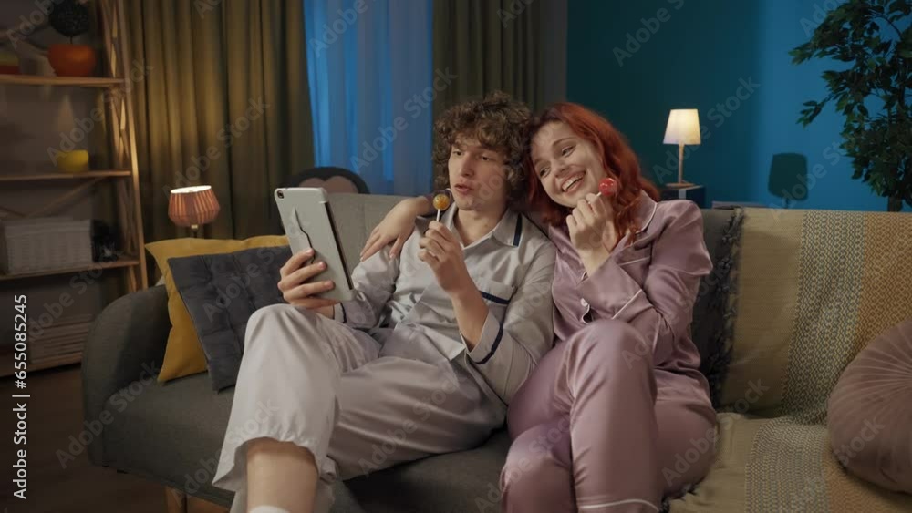 Portrait of young couple spending time together. Man and woman in pajamas on the sofa eating lollipops, talking by video call on tablet. Portrait of young couple spending time together. Man and woman in pajamas on the sofa eating lollipops, talking by video call on tablet.