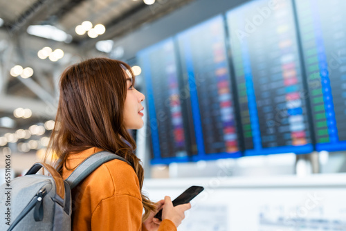 Young asian woman in international airport, using mobile smartphone and checking flight at the flight information board