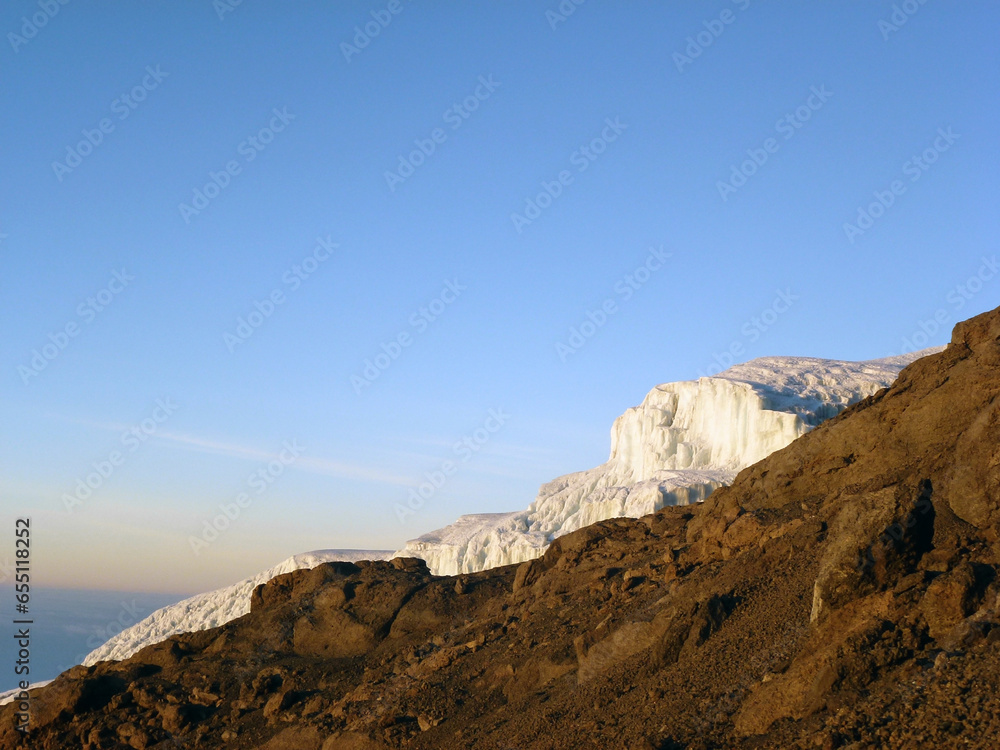 A rocky mountain slope behind which is an iceberg on the background of a blue sky