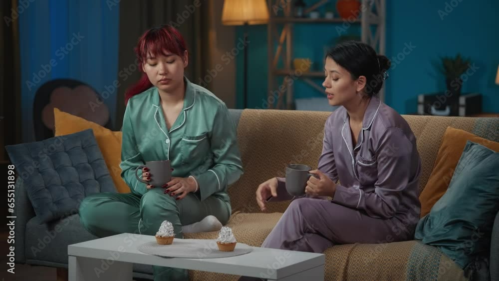 Medium-full video capturing two young women wearing pajamas sitting on the couch, talking about something, drinking tea of coffee and eating cupcakes, messing around. Medium-full video capturing two young women wearing pajamas sitting on the couch, talking about something, drinking tea of coffee and eating cupcakes, messing around.