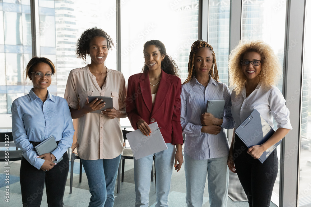Corporate portrait smiling successful African American businesswomen team standing in office room with panoramic windows, happy confident employees colleagues staff members looking at camera Corporate portrait smiling successful African American businesswomen team standing in office room with panoramic windows, happy confident employees colleagues staff members looking at camera