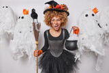 Photo of angry mysterious sorceress prepares for halloween carnival wears black clothing holds broom and cauldron frowns face from annoyance surrounded by scary ghosts. Autumn holiday concept
