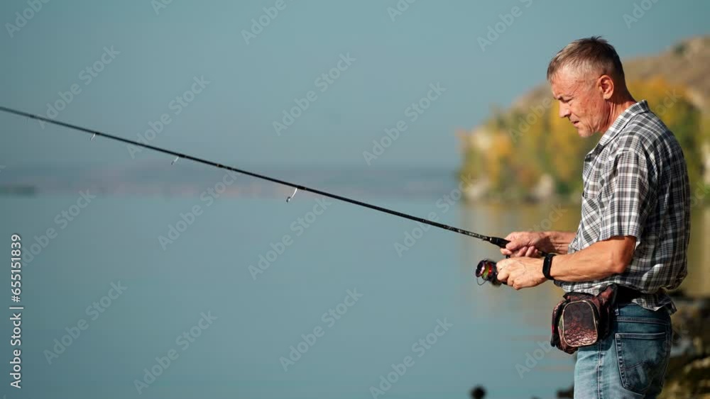 In autumn, a mature adult man in a shirt and jeans catches fish with a spinning rod on the river bank. Senior adult goes fishing on weekends. A young pensioner is engaged in a hobby. In autumn, a mature adult man in a shirt and jeans catches fish with a spinning rod on the river bank. Senior adult goes fishing on weekends. A young pensioner is engaged in a hobby.