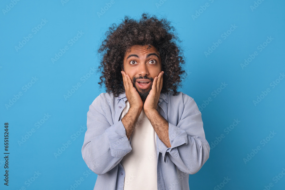 Young surprised optimistic Arabian man puts hands on cheeks and opens mouth having experienced pleasant shock from received offer or unexpected news about career advancement stands in blue studio. Young surprised optimistic Arabian man puts hands on cheeks and opens mouth having experienced pleasant shock from received offer or unexpected news about career advancement stands in blue studio.