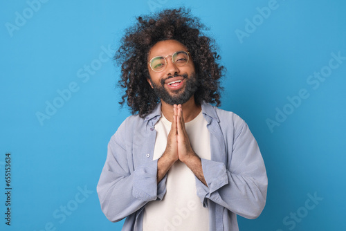 Young kind positive Arabian man smiles and makes prayer gesture demonstrating readiness to forgive offender committed unpleasant act dressed in casual style stands on blue studio background.