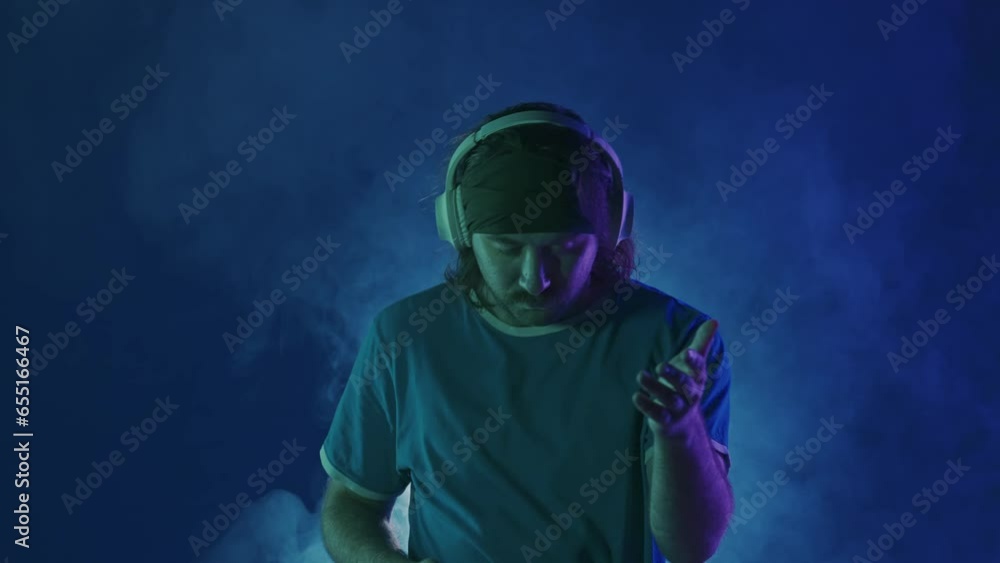 A man wearing wireless headphones dances while enjoying the music. A man dances in a studio under a spotlight, in clouds of smoke, in pink and green neon lights. A man wearing wireless headphones dances while enjoying the music. A man dances in a studio under a spotlight, in clouds of smoke, in pink and green neon lights.