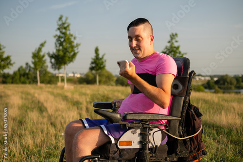 Man wearing pink t-shirt in wheelchair holds smartphone against countryside view. Male with hands disability talking to friend on videocall