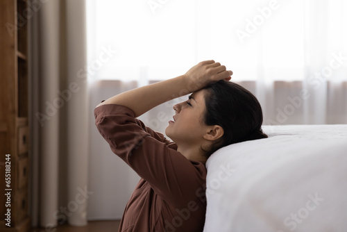 Side view exhausted unhappy Indian woman touching forehead, feeling strong headache, unhealthy tired young female suffering from migraine, thinking about problems, sitting on floor at home alone