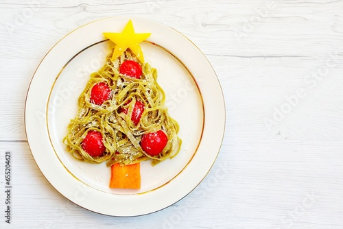 Christmas tree pasta made it from spinach tagliatelle pasta,tomatoes,carrot,parmesan cheeses and yellow bell peppers on plate with white wood background.Creative art food idea for Christmas's dinner


