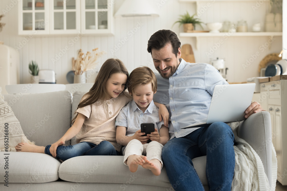 Happy caring father spends time with children at home, cute kids use mobile phone for entertainment watch funny videos on social media, dad controls internet usage. Two generations enjoy modern tech