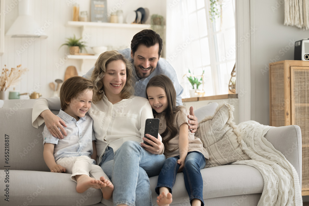 Happy family spend time in studio apartment sit on couch, smiling mother hold mobile phone take selfie photo together for memory, kids have great time with parents, make online call with grandparents Happy family spend time in studio apartment sit on couch, smiling mother hold mobile phone take selfie photo together for memory, kids have great time with parents, make online call with grandparents