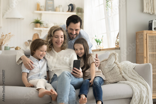 Happy family spend time in studio apartment sit on couch, smiling mother hold mobile phone take selfie photo together for memory, kids have great time with parents, make online call with grandparents