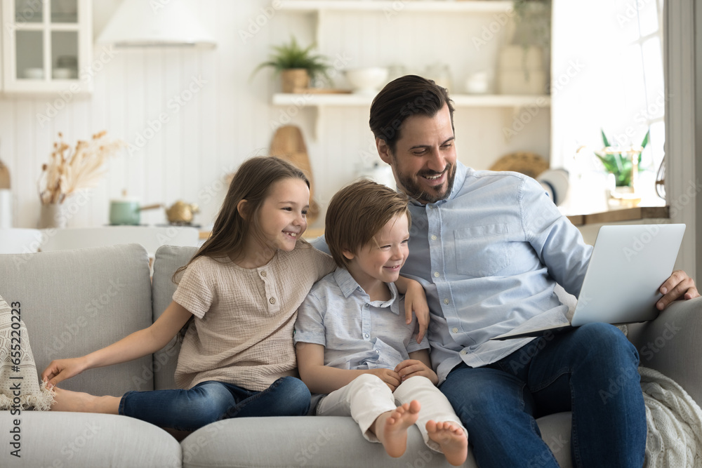 Happy handsome young father sits on sofa enjoy weekend time with cute little 6s children, hold laptop watch funny video, online TV family channel, doing e-shopping, laugh feels happy, use modern tech Happy handsome young father sits on sofa enjoy weekend time with cute little 6s children, hold laptop watch funny video, online TV family channel, doing e-shopping, laugh feels happy, use modern tech