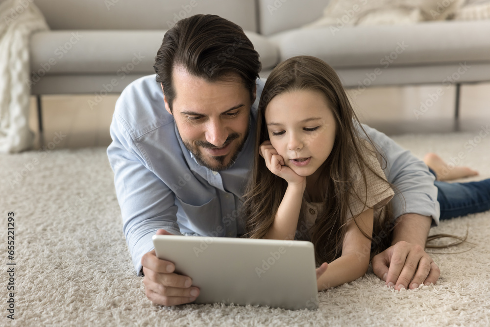 Fototapeta premium Smiling father and daughter choose funny movie on digital tablet lying on floor in living room, dad show new modern app to daughter, watch educational video, enjoy friendly relations, rest together