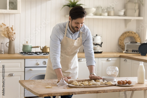 Handsome young man wear apron flattening dough with rolling pin stand at dining table in modern kitchen, make pastry or dumplings, prepare delicious food snack for family, enjoy culinary hobby at home