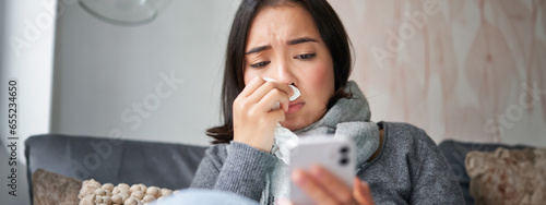 Obraz na płótnie Ill korean woman sitting at home, sneezing, caught cold, reading news on mobile