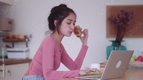 Young happy Asian Freelance woman eating croissant for breakfast and drinking coffee while working with laptop on kitchen counter, enjoys working from home