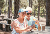 Cheerful senior couple enjoying a sandwich while sitting at a wooden table in the woods using mobile phone in video chat. Happy elderly man and woman appreciating nature and freedom during retirement