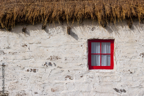 Old white weathered thatched roof cottage with a red square window