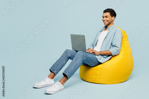 Full body young IT man of African American ethnicity wear shirt casual clothes sit in bag chair hold use work on laptop pc computer isolated on plain pastel light blue cyan background studio portrait.