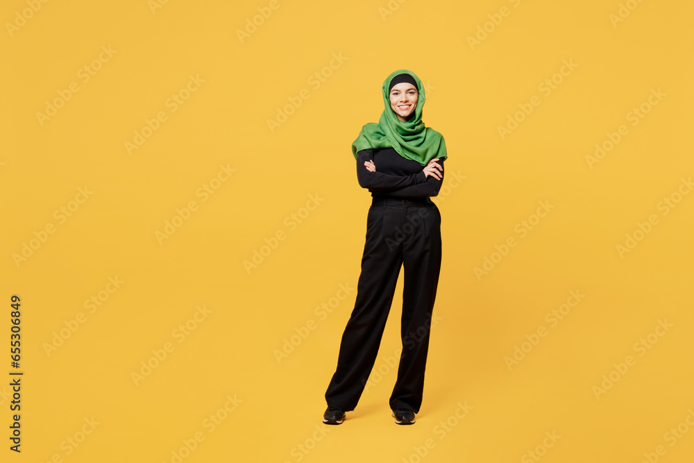 Full body smiling young arabian asian muslim woman wear green hijab abaya black clothes hold hands crossed folded look camera isolated on plain yellow background. People uae islam religious concept. Full body smiling young arabian asian muslim woman wear green hijab abaya black clothes hold hands crossed folded look camera isolated on plain yellow background. People uae islam religious concept.