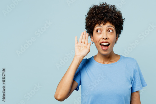 Young curious nosy woman of African American ethnicity wear t-shirt casual clothes try to hear you overhear listening intently isolated on plain pastel light blue cyan background. Lifestyle concept.