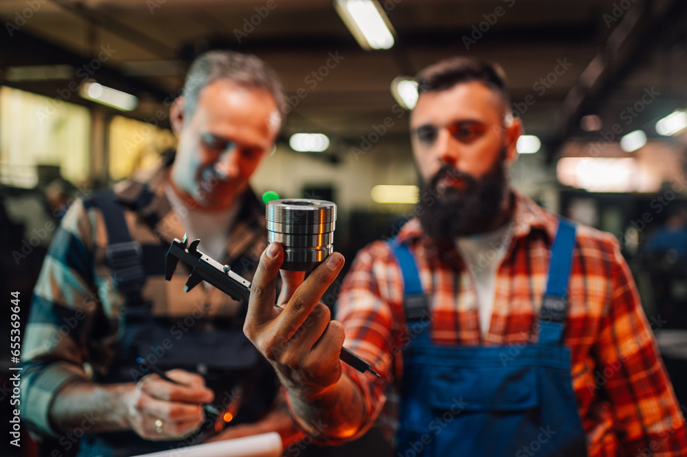 Focus frame of a hand holding a caliper and a metal piece, two people blurred in background. Focus frame of a hand holding a caliper and a metal piece, two people blurred in background.