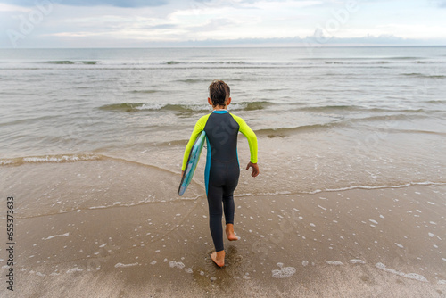 Young boy in wet suit at the beach running into the water while carrying his boogie board