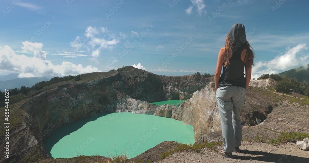 Naklejka premium Tourist woman enjoy morning view on volcanic Kelimutu blue crater lakes. Nature background. Travel destination. Beautiful wild landscape. Exotic summer vacation, hiking, relax. Close up back view