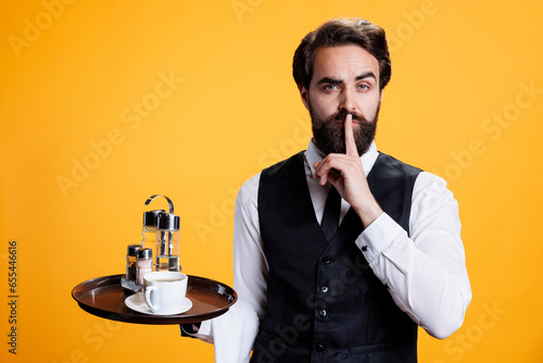 Fotografi Secretive waiter doing mute symbol in studio, presenting privacy and confidential gesture with finger over mouth