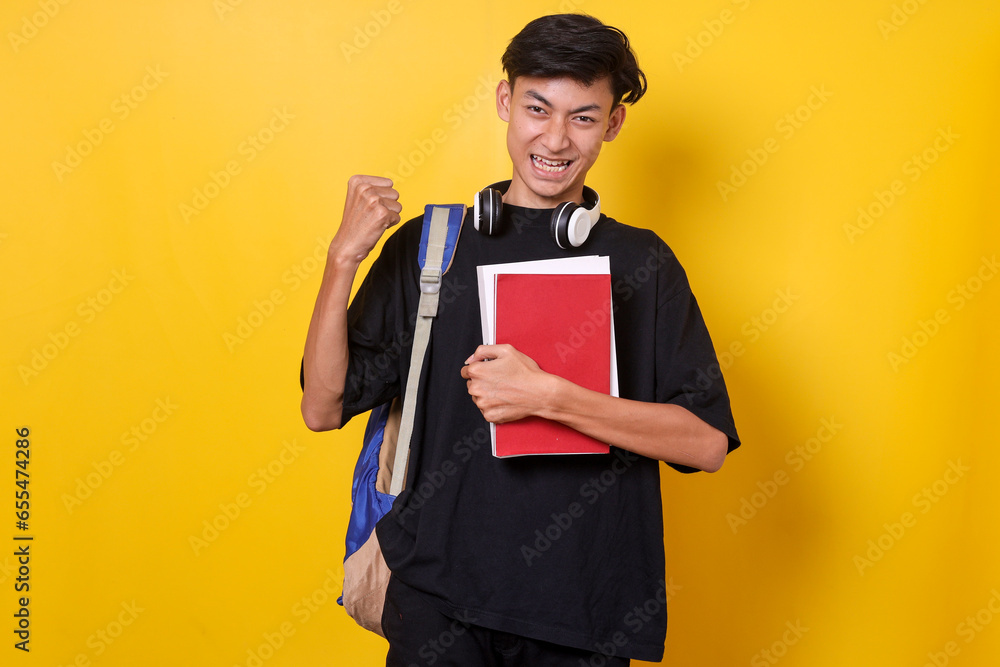 Excited male student wearing backpack and hold books showing winner expression celebrating victory over yellow background Excited male student wearing backpack and hold books showing winner expression celebrating victory over yellow background