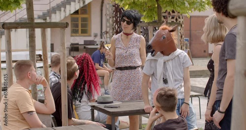 Young woman holding glass of non-alcoholic beverage joins group of young people. Her appearance is unconventional for teacher, reflecting sense of freedom in self-expression. Man wearing a horse mask photo