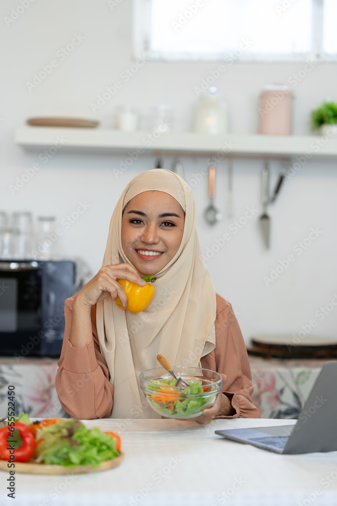 A young Muslim woman in a hijab holds bell peppers while preparing a salad to nourish her skin. While using my laptop to research menus for cooking healthy vegan food ideas. A young Muslim woman in a hijab holds bell peppers while preparing a salad to nourish her skin. While using my laptop to research menus for cooking healthy vegan food ideas.
