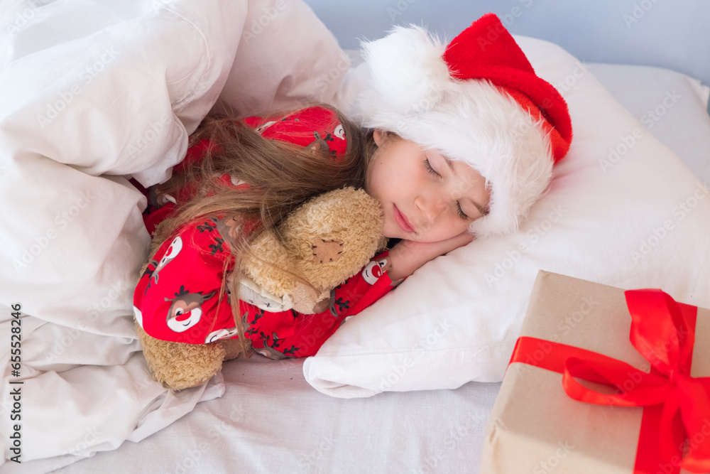 Merry Christmas and happy holidays. A cute little girl of 6-8 years old in a Santa hat is sleeping at home in bed hugging a Teddy toy, there is a gift box next to it.
