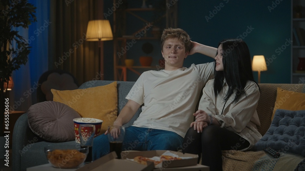 Portrait of teenage couple spending leisure time. Boy and girl sitting on the sofa eating popcorn and watching something on tv, boy tries to hug the girl.