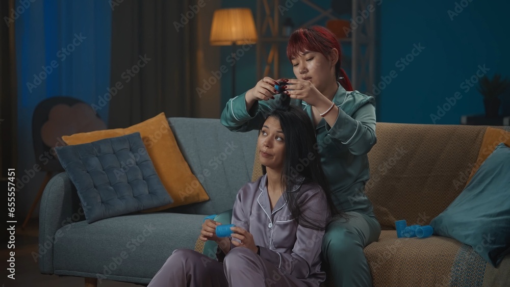 Naklejka premium Medium-full photo capturing two young women wearing pajamas sitting on the couch, gossiping, talking about something as one of them is doing the other one's hair.