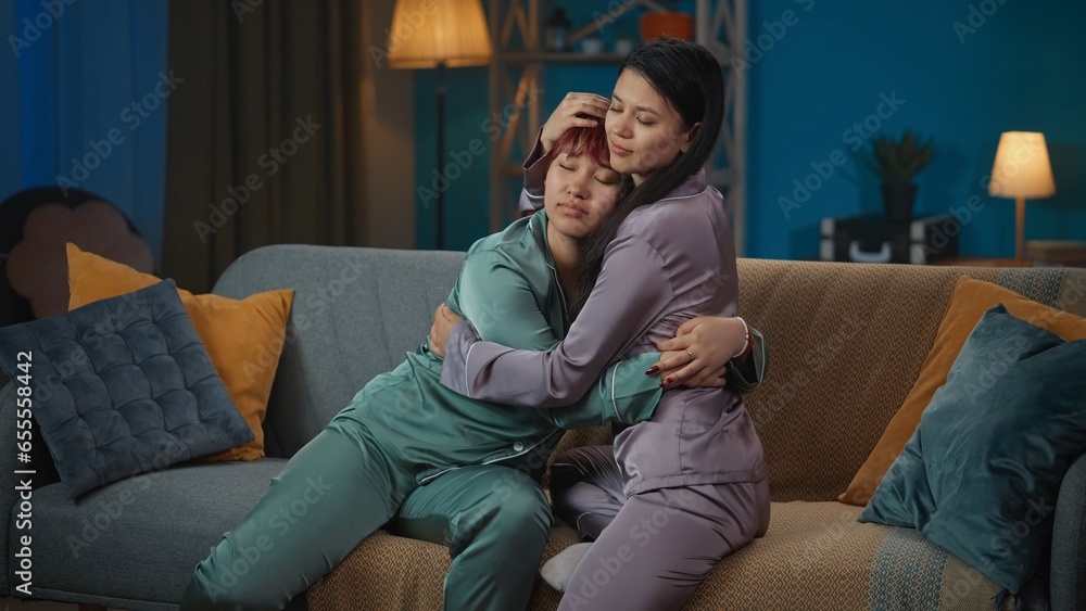 Medium-full photo capturing two young women wearing pajamas sitting on the couch in a room, hugging. They made up after a fight.