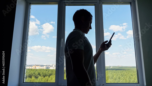 Male office worker standing near window scrolls social media on smartphone. Man employee silhouette in t-shirt reads news on Internet on smartphone.