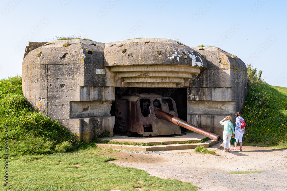 Two tourists observe the 150 mm gun in a bunker in the Longues-sur-Mer battery in Normandy, France, a WWII German coastal artillery battery part of the Atlantic Wall fortifications. - obrazy, fototapety, plakaty 