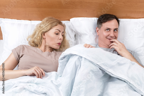 Two in bed, a woman looks with interest under the blanket, a man smiles maliciously. Relationship concept photo