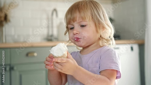 Baby girl enjoying ice cream. Pretty little toddler eating an ice-cream indoors, at home. Dining room background. Small child eats plombir and cream messy on her mouth. Cute kid with tasty sweet food. photo