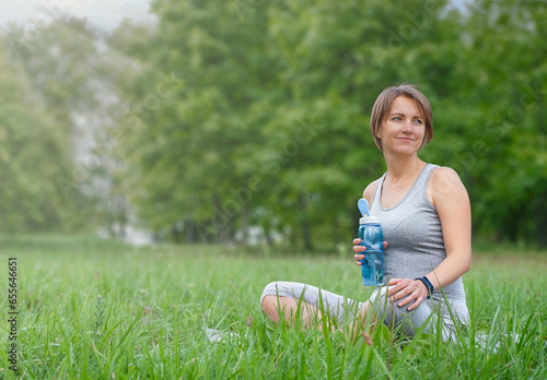 Woman with bottle of water rests after yoga exercises outdoor in natural park. Active lifestyle concept