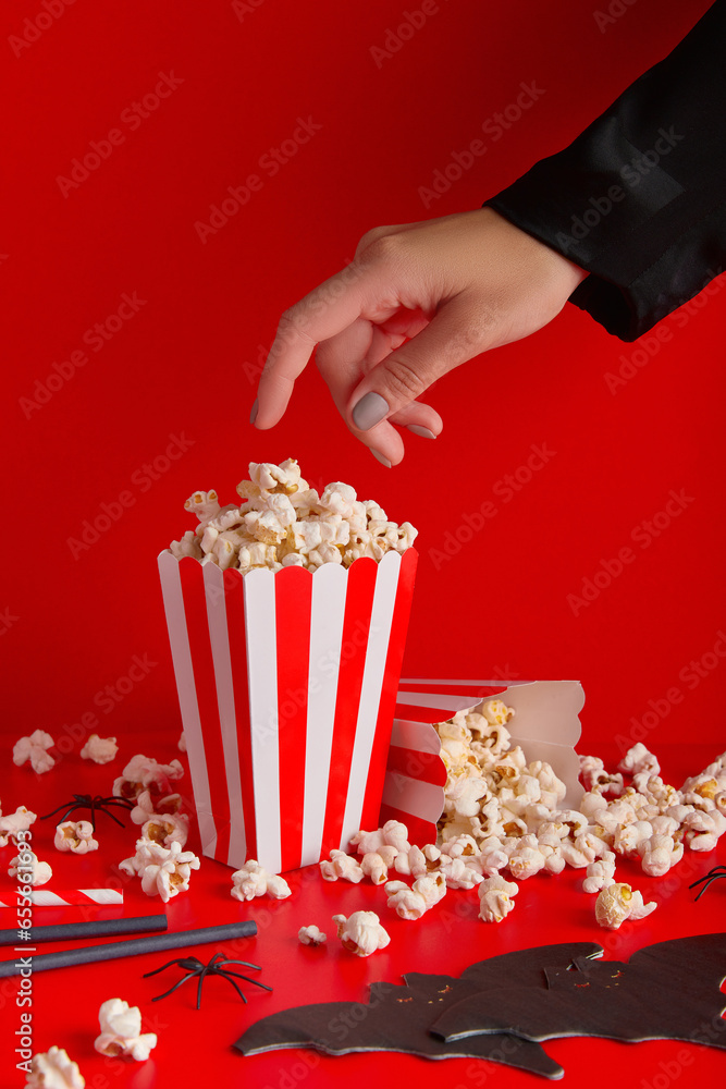 Womans hand taking popcorn from striped box, bat napkin and straws on a red background.