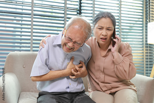 Asian senior man lying down and suffering from chest pain or heart attack from working accident on sofa in living room. Elderly woman caregiver consoling and help while hugging support and first aid