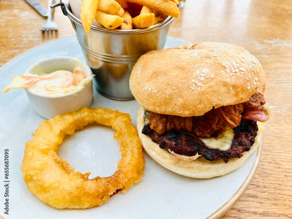 Beef burger in a white bread roll with French fries, onion ring and coleslaw served on a white plate