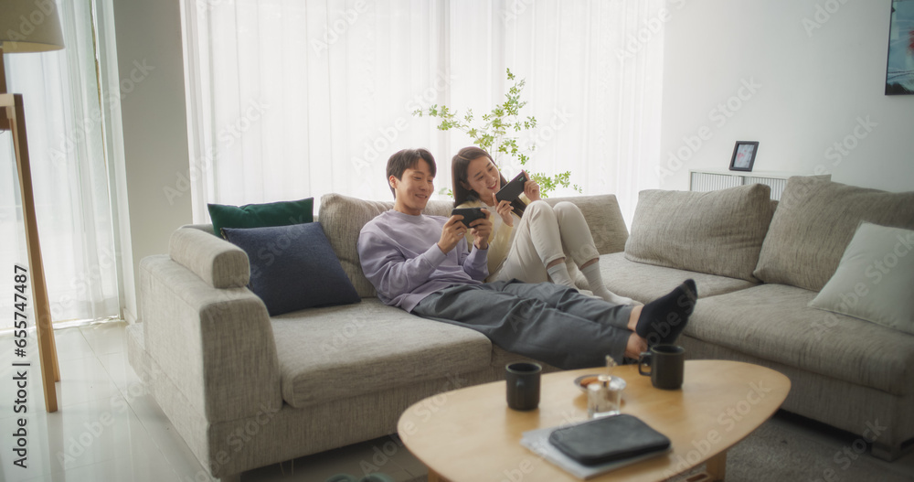 South Korean Couple Sitting on a Cozy Couch, Playing Funny Arcade Video Games Together in a Stylish Bright Living Room. Beautiful Couple in a Relationship Enjoying Time on a Sofa at Home