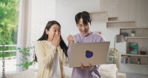 Young and Lovely South Korean Couple Standing in a Stylish Living Room and Using Laptop Computer at Home. Man and Woman Waiting for Successful Results, Happy, Cheering and Celebrating the Win