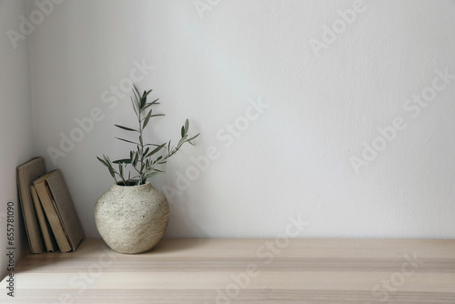 Moody Mediterranean home design. Vintage vase with olive tree branches. Old books on wooden table. Living room still life, home office. Empty white wall copy space. Modern interior, no people.