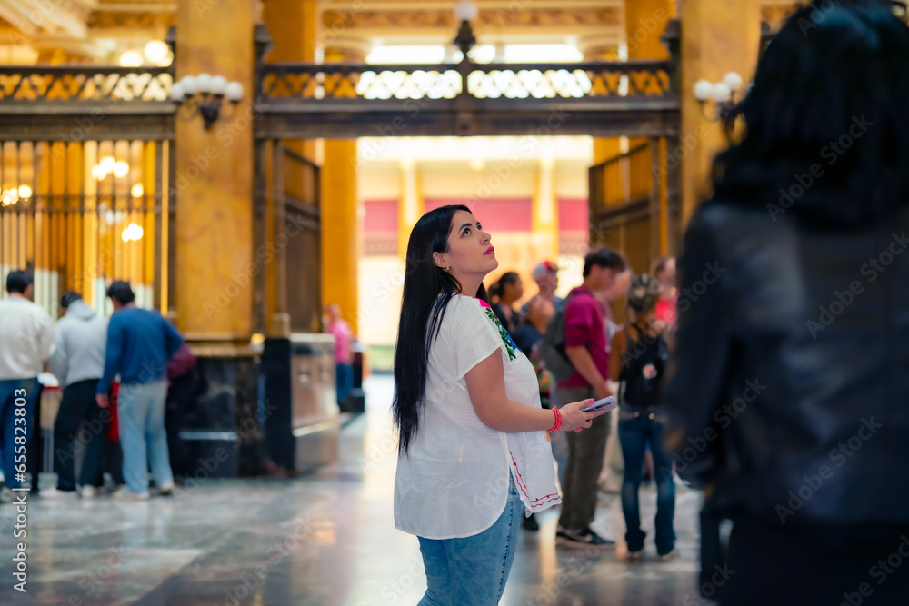 Happy young mexican woman tourist in embroidered top looking around while standing in Postal palace in downtown Mexico City and in light against blurred interior - obrazy, fototapety, plakaty 