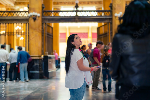Happy young mexican woman tourist in embroidered top looking around while standing in Postal palace in downtown Mexico City and in light against blurred interior photo